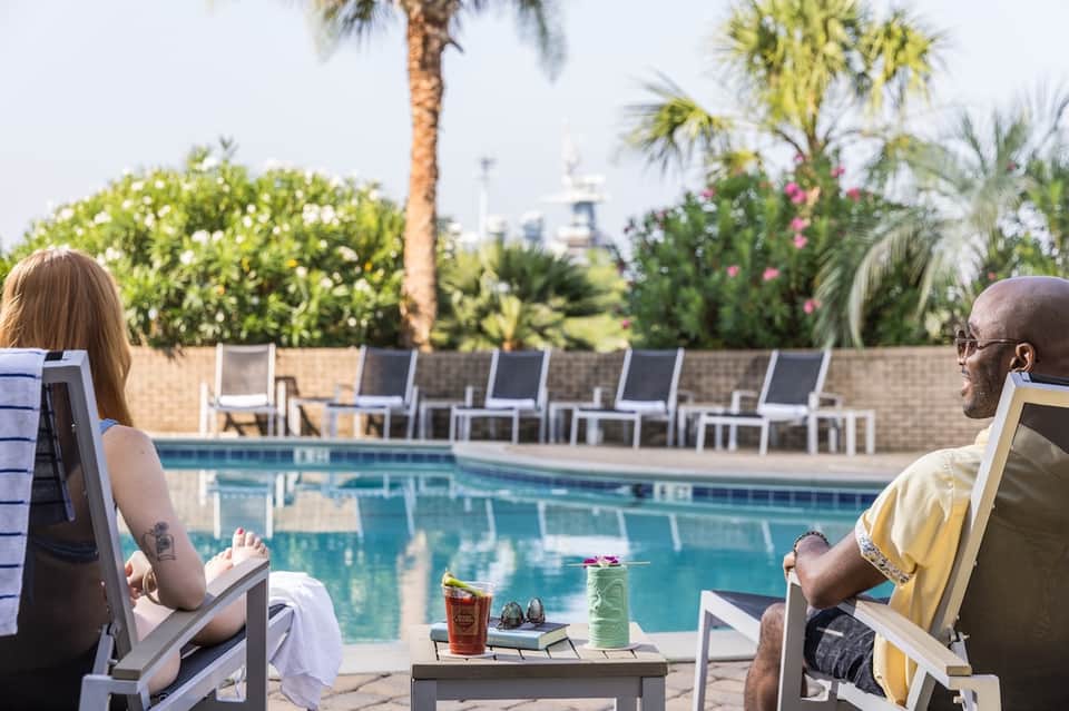 Couple relaxing poolside on lounge chairs with drinks and palm trees in background