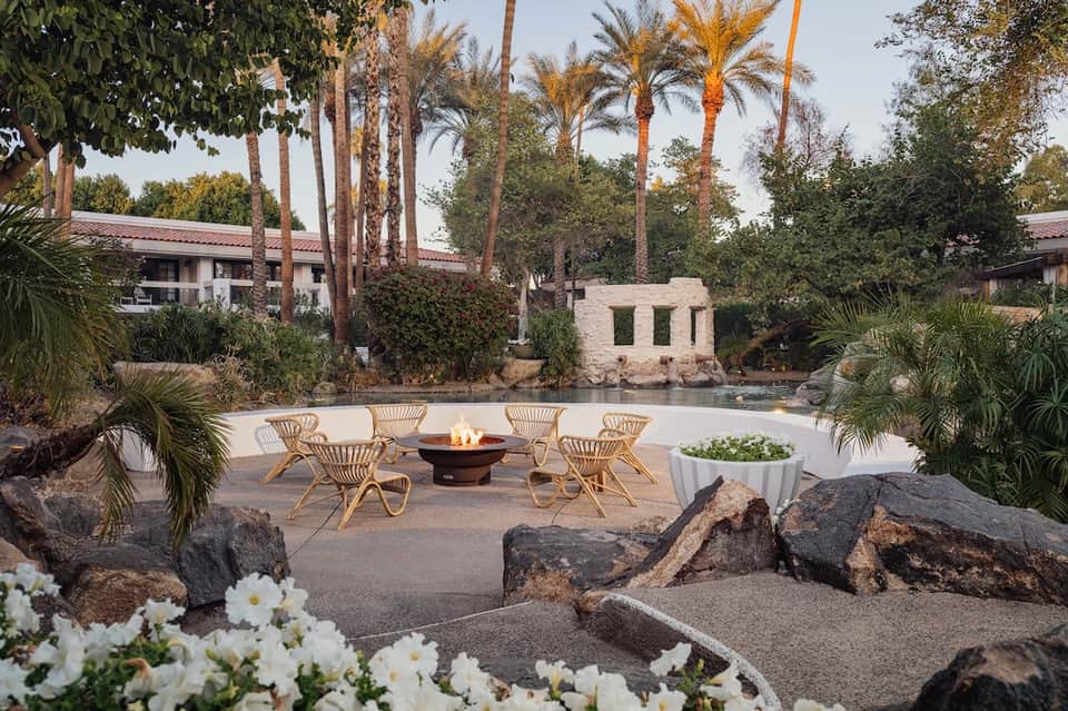 Outdoor fire pit lounge with woven chairs, palm trees, water feature, and tropical landscaping