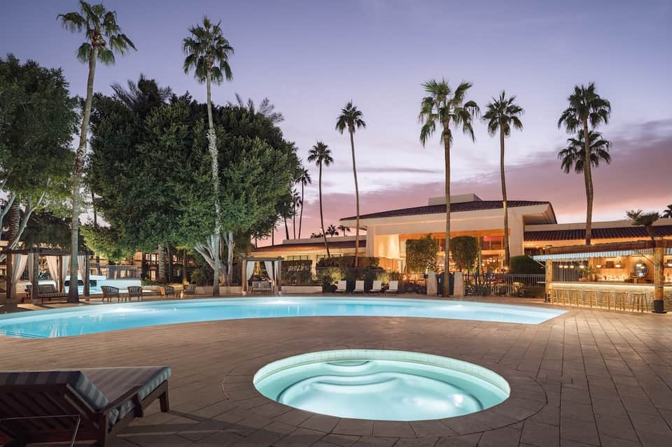 Resort pool and spa at dusk with palm trees, illuminated buildings, and hot tub in foreground
