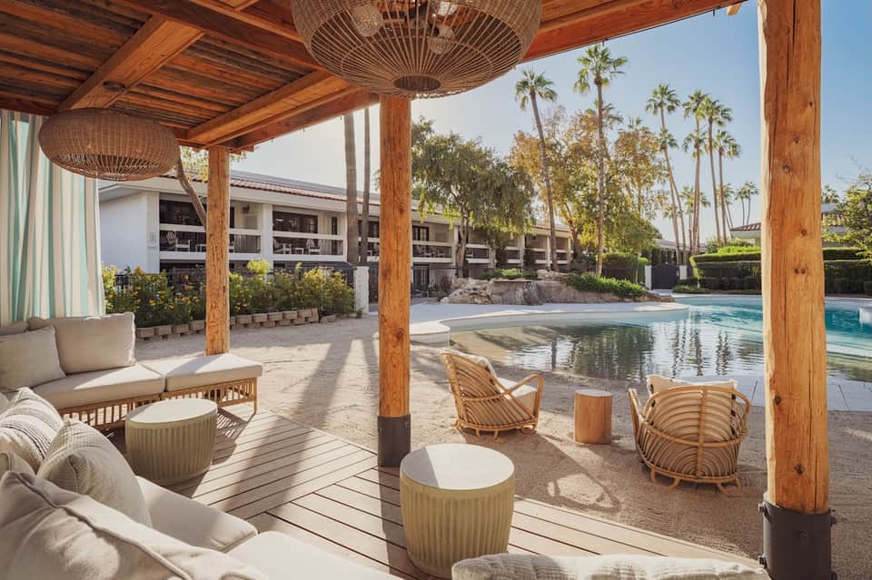 Poolside covered patio with wooden pergola, woven pendant lights, lounge seating, and resort buildings with palm trees