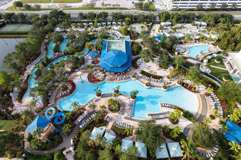 Aerial view of sprawling resort with multiple pools, blue umbrellas, water features, and landscaping