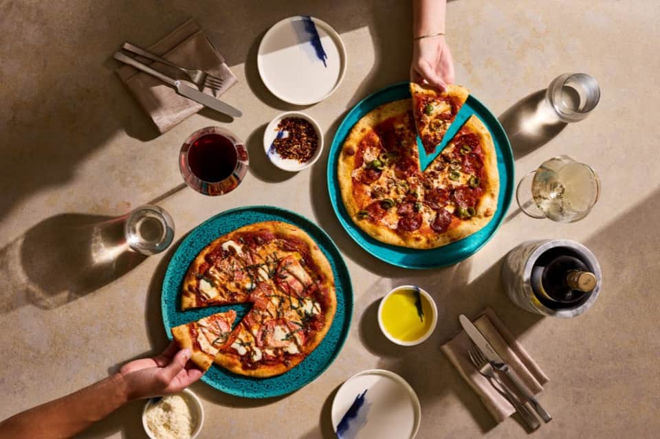 Overhead view of two pizzas on turquoise plates with wine, water, and place settings on beige table