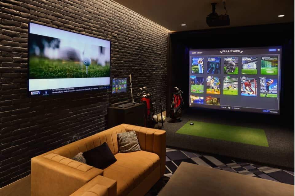 Game room and sports lounge with golf simulator, large screen displays, orange sofa, brick walls, and gaming equipment