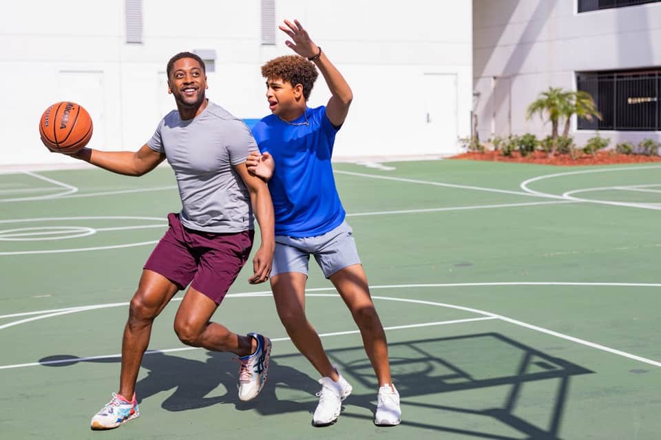 Two men playing basketball on an outdoor court in front of a white building