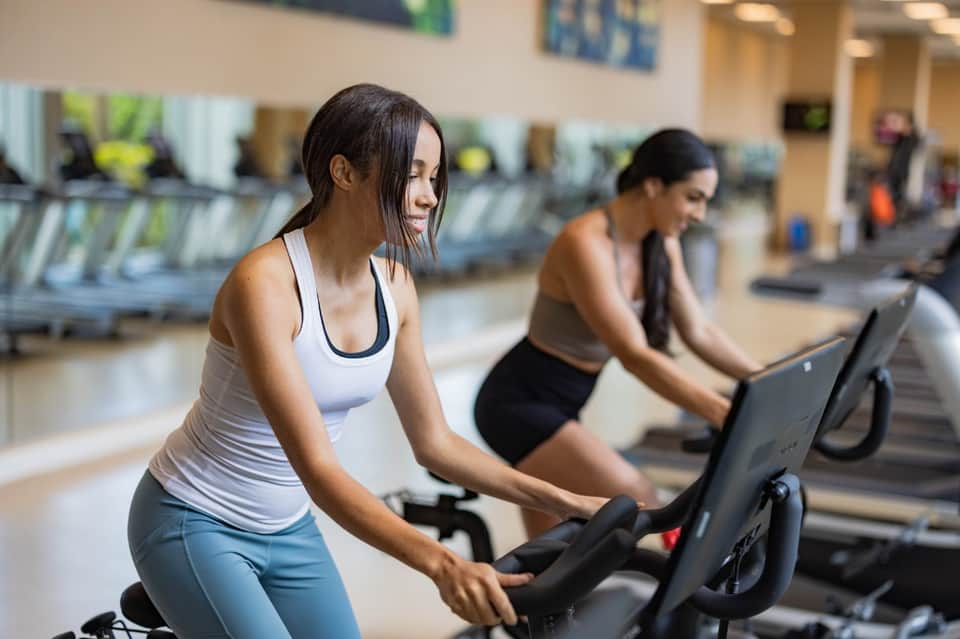Women on stationary bikes in bright fitness center with rows of equipment