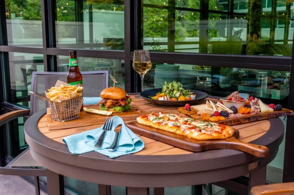 Modern restaurant table with burger, fries, pizza, salad, charcuterie board, wine glass, and green outdoor views