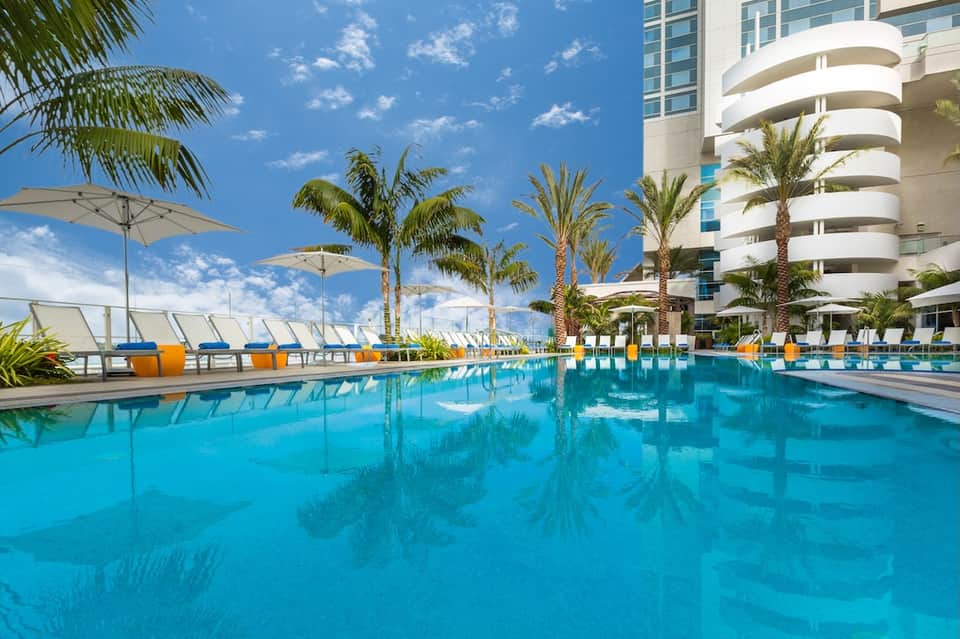 Resort pool with crystal blue water, palm trees, white umbrellas, and modern curved architecture