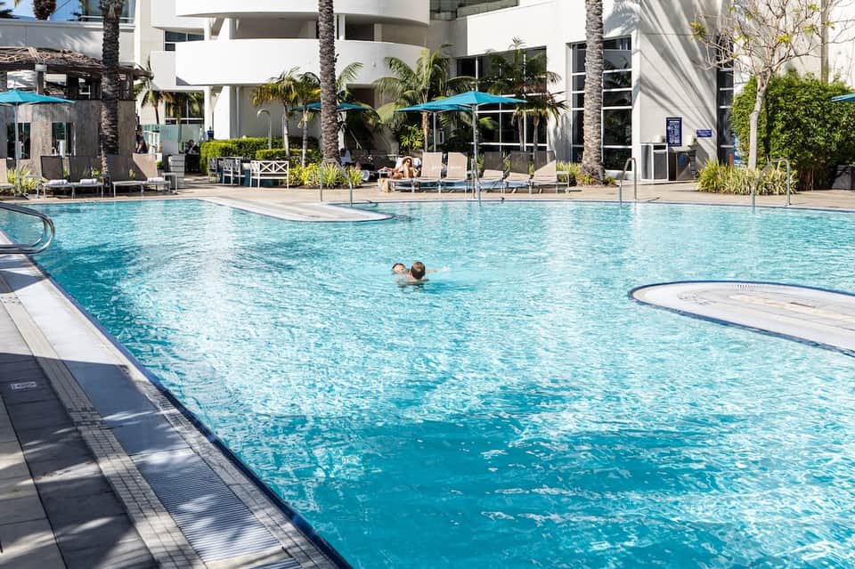 Resort pool with swimmers, white modern building, palm trees, and blue umbrellas