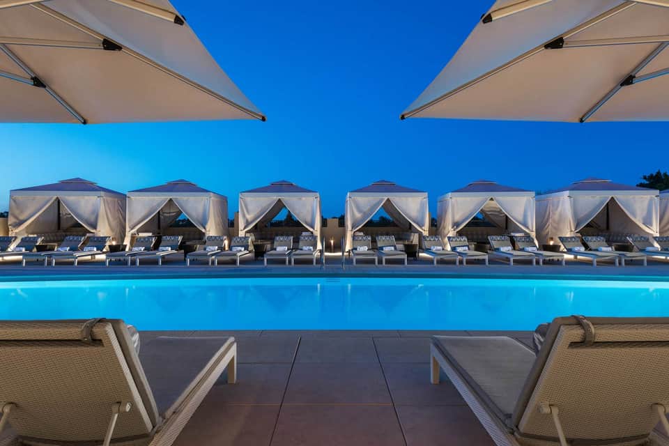 Modern resort pool at dusk with white cabanas and lounge chairs under clear blue sky