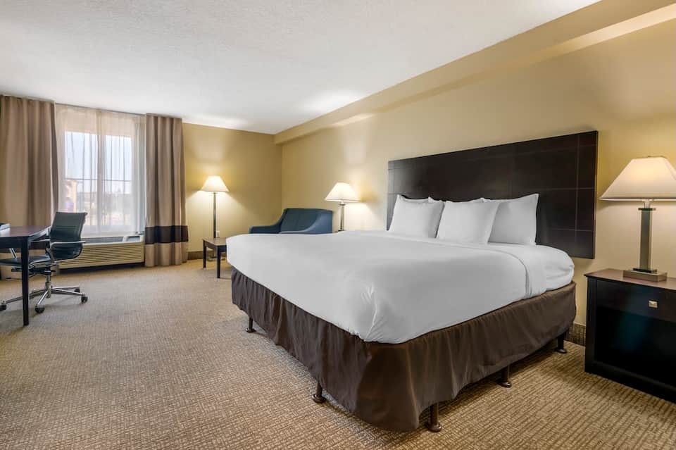Spacious hotel bedroom with queen bed, black headboard, beige walls, work desk, and window with city views.