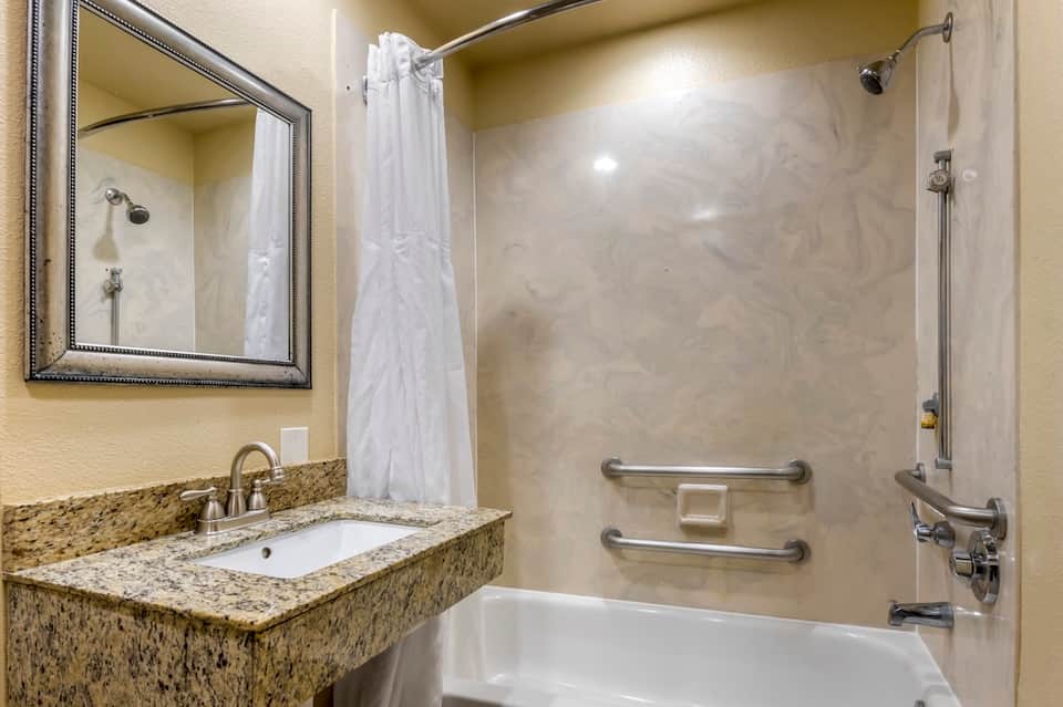 Hotel bathroom with granite countertop sink, mirror, white shower curtain, and tub with grab bars