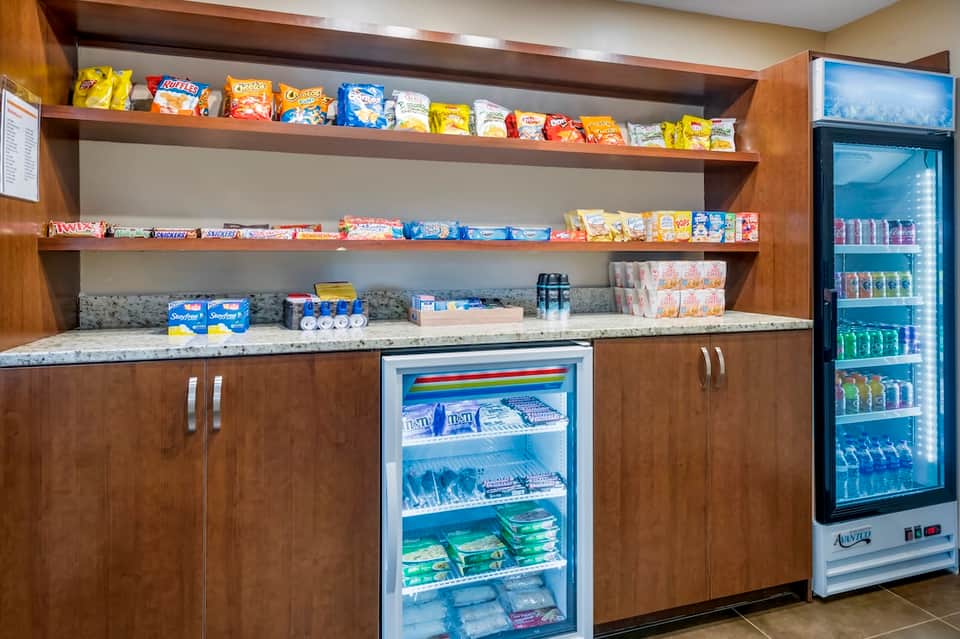 Hotel snack bar with wooden cabinetry, open shelving displaying candies and snacks, and beverage cooler.