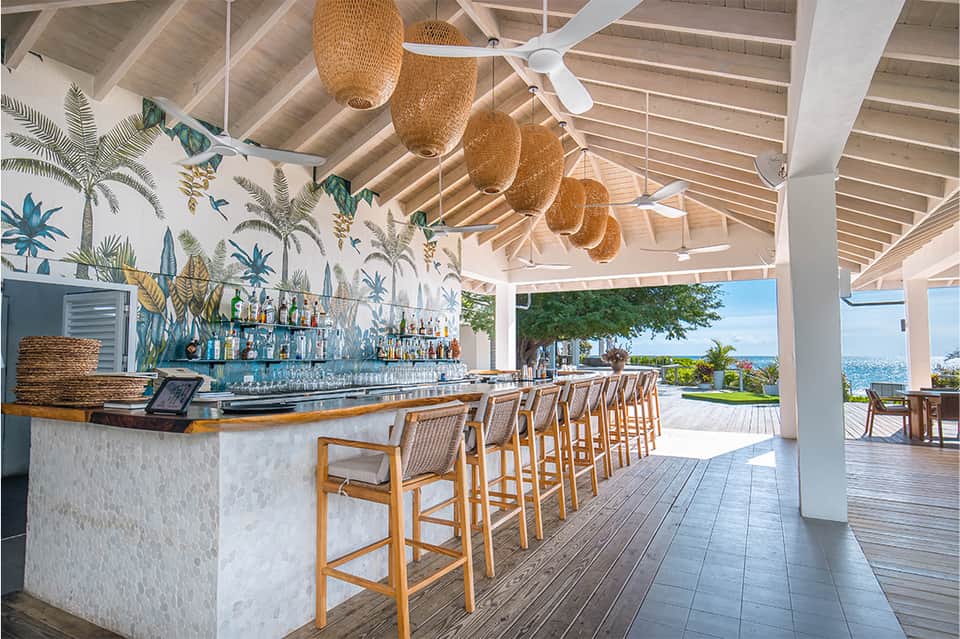 Tropical beach bar with white columns, woven pendant lights, wooden stools, and ocean views