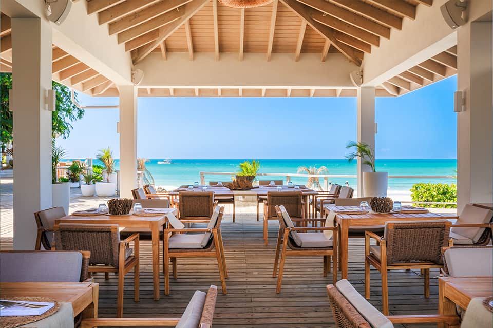 Beachfront dining pavilion with wooden tables, rattan chairs, and clear ocean view under white roof