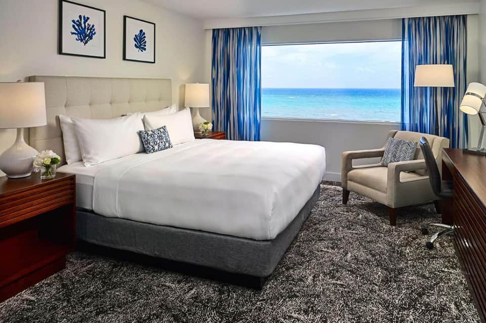Oceanfront hotel bedroom with beige headboard, white bedding, blue curtains, and beach view