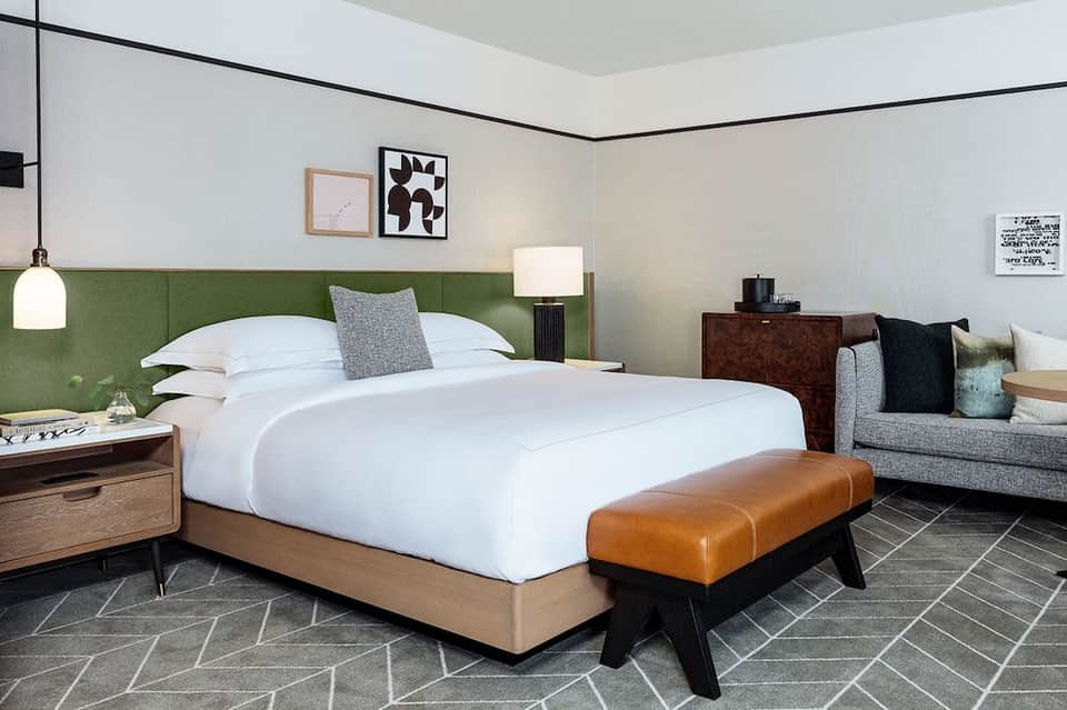 Contemporary hotel bedroom with green headboard, white bedding, wood furniture, and modern artwork.