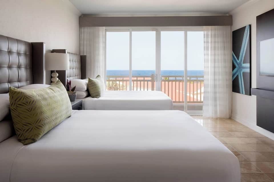 Oceanview hotel bedroom with two queen beds, balcony doors, and sea horizon visible in distance