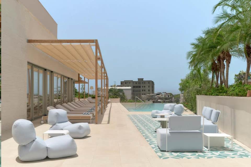 Rooftop terrace with modern white seating, wooden pergola, pool, and palm trees against city skyline