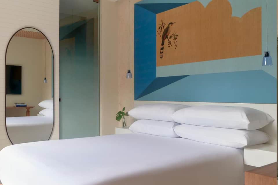Modern hotel bedroom with white bedding, arched mirror, blue and tan wall mural, and pendant lighting