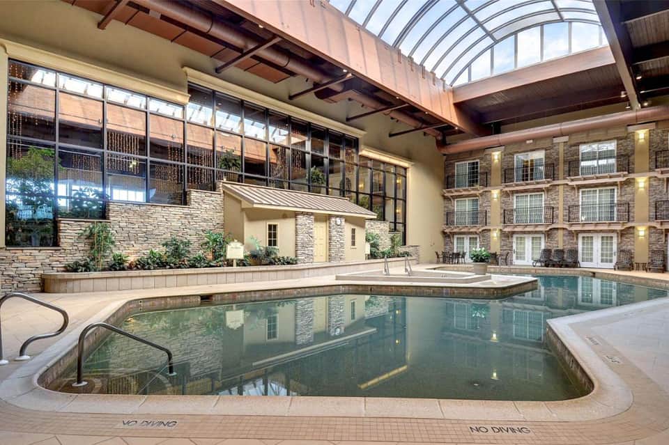 Indoor heated swimming pool with stone walls, arched glass ceiling, and multi-story hotel rooms