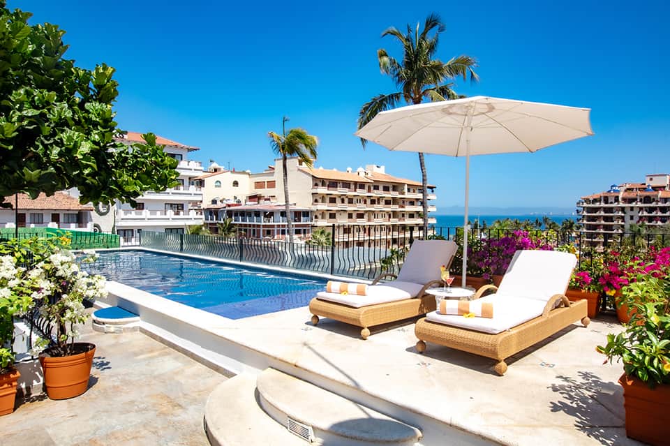 Mediterranean resort pool with white umbrella, lounge chairs, sea view, and colorful blooming flowers