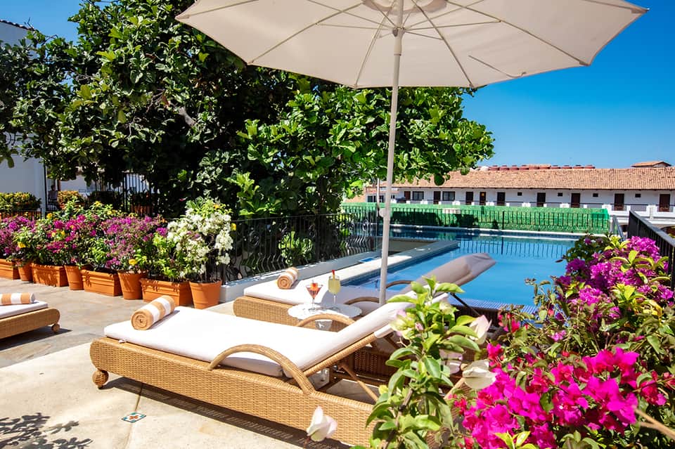 Poolside lounge area with chaise lounges, white umbrella, vibrant magenta flowers, and water view