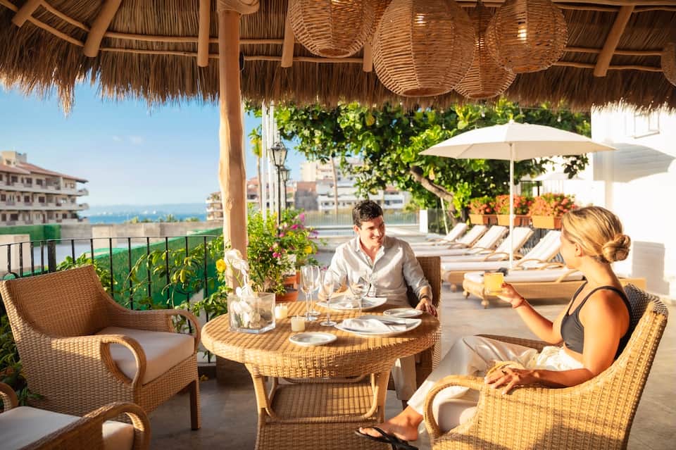 Couple dining at a round table on a rooftop terrace with ocean views and lounge chairs in background
