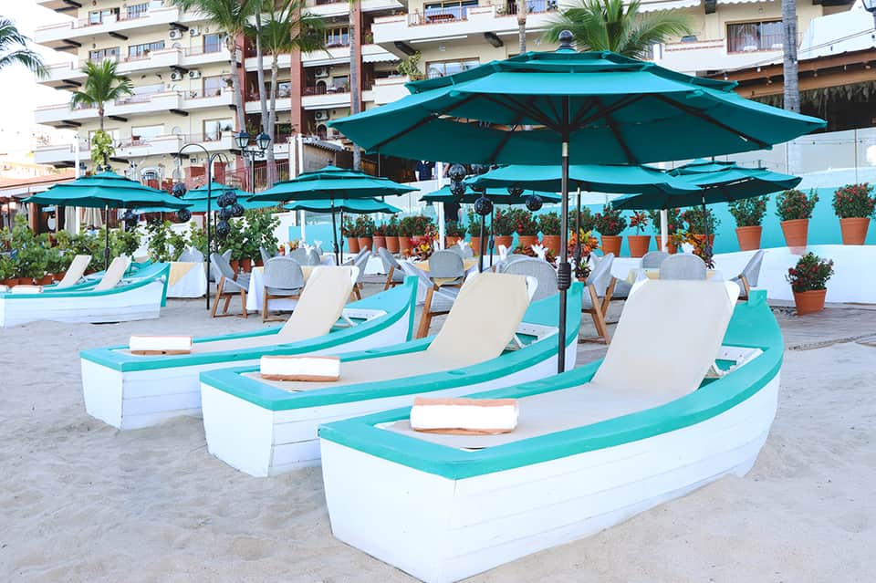 Beach loungers with turquoise umbrellas and cushions arranged on sand near resort building