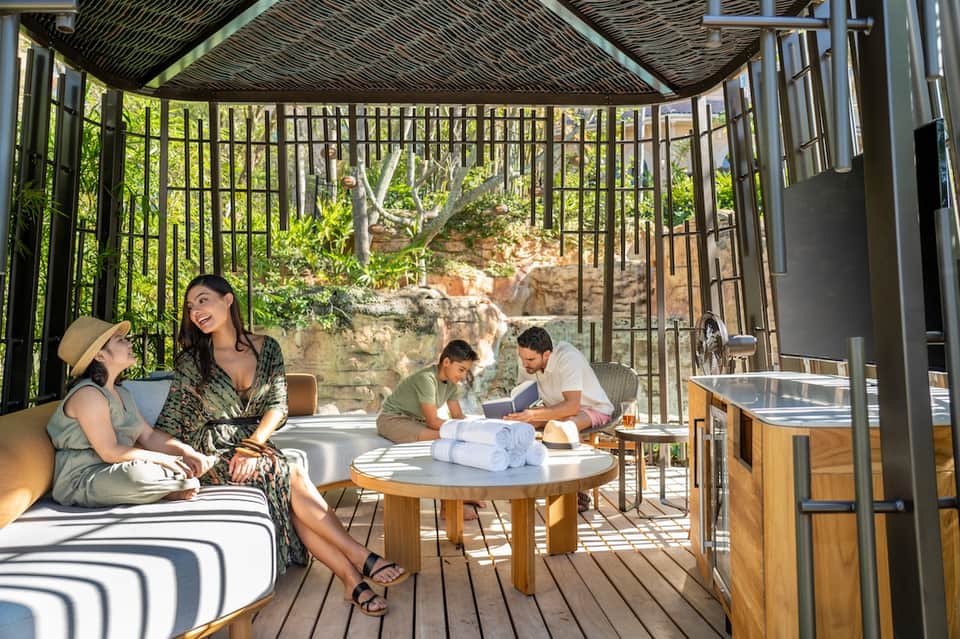 Family relaxing on covered patio with wooden furniture, green metal railings, and stone architecture