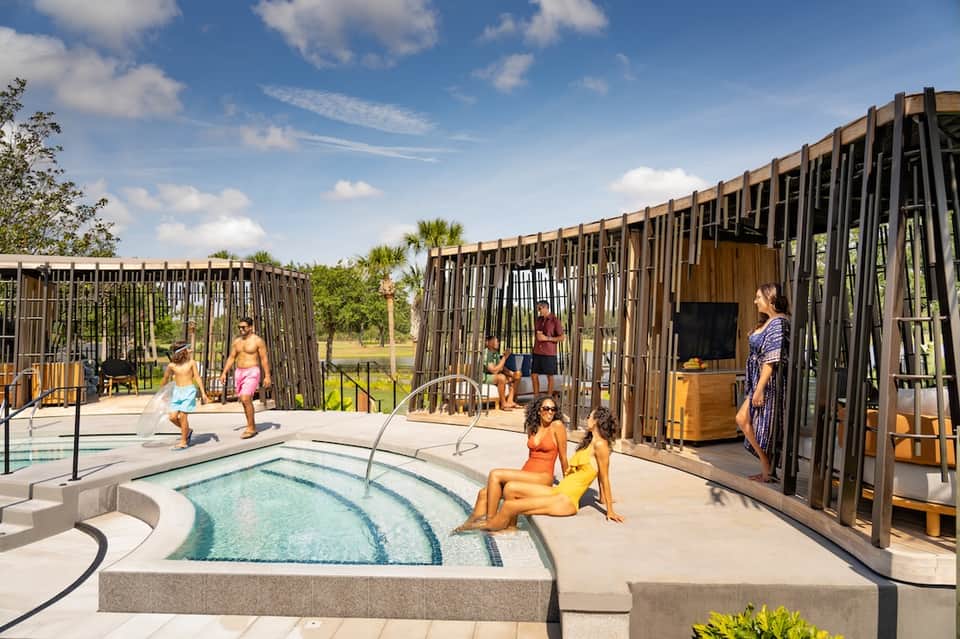 Resort pool area with hot tub, families enjoying water activities, modern wooden cabanas, palm trees, lake view