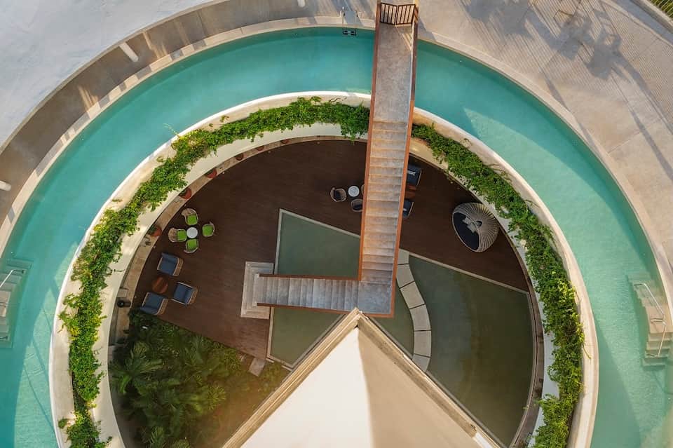 Aerial view of circular modern building with turquoise pool surrounding a central structure with wooden deck and green plantings