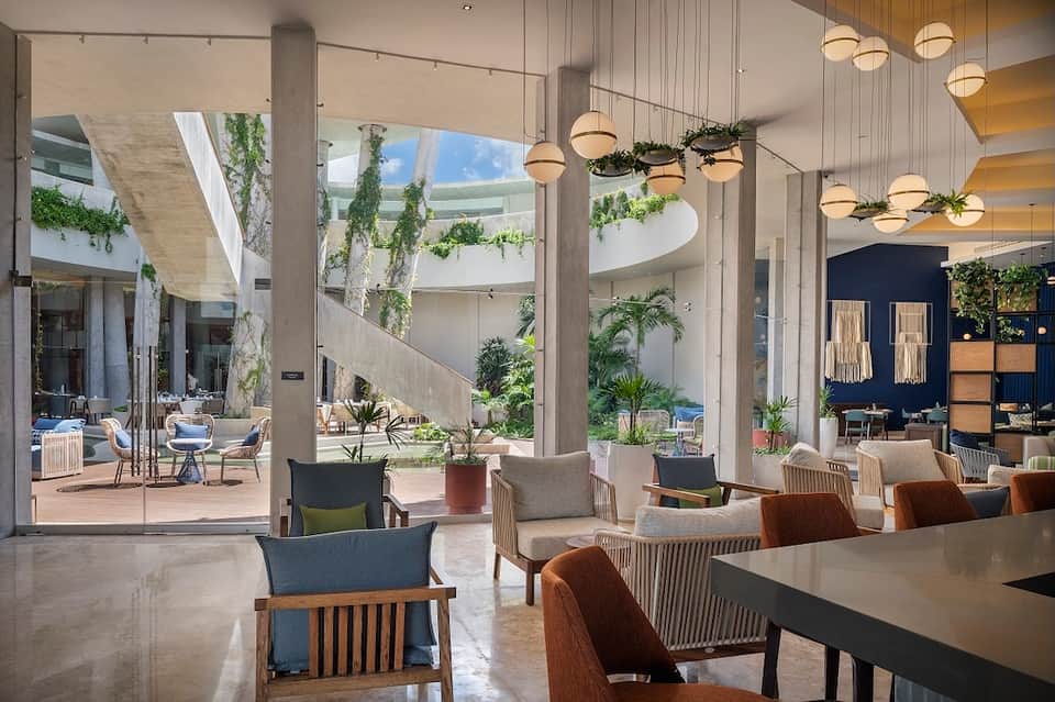 Modern hotel lobby with concrete columns, hanging plants, pendant lights, and ocean view through glass doors