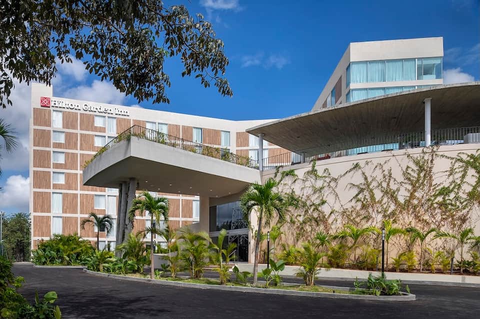 Contemporary hotel exterior with wood-paneled facade, overhanging concrete balcony, and tropical plantings