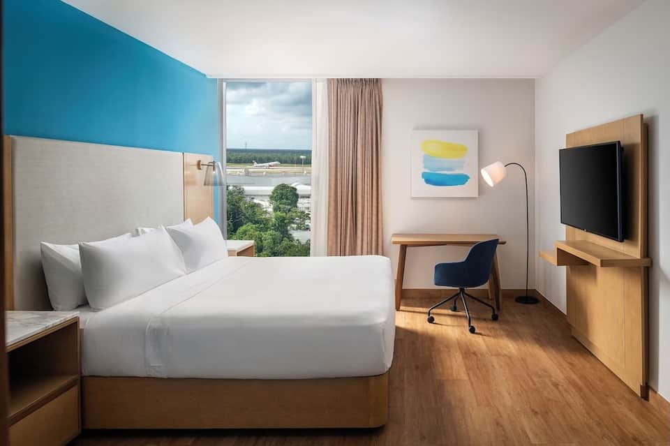 Hotel bedroom with blue accent wall, queen bed, wooden desk, TV, and panoramic window overlooking river landscape
