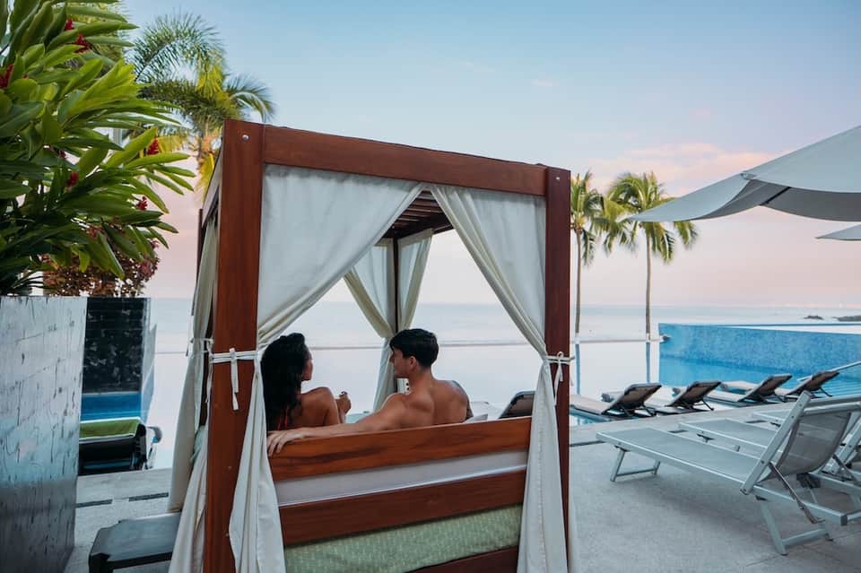 Couple relaxing in wooden cabana bed overlooking infinity pool and ocean horizon at dusk
