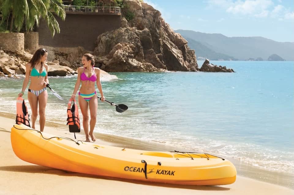 Two women in bikinis with kayaking gear standing on a beach near an orange ocean kayak