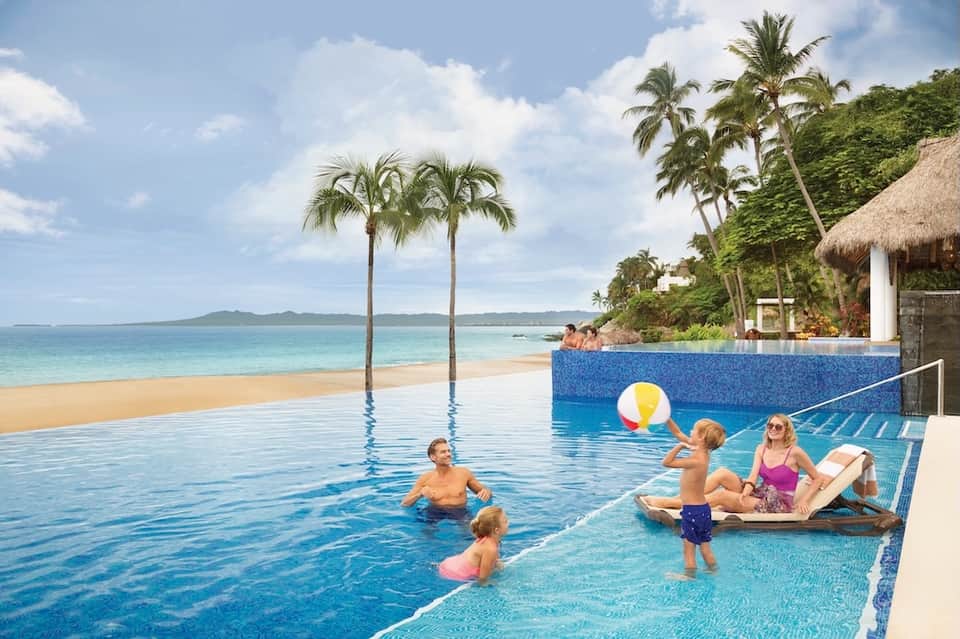 Beachfront infinity pool with palm trees, ocean view, families swimming and relaxing
