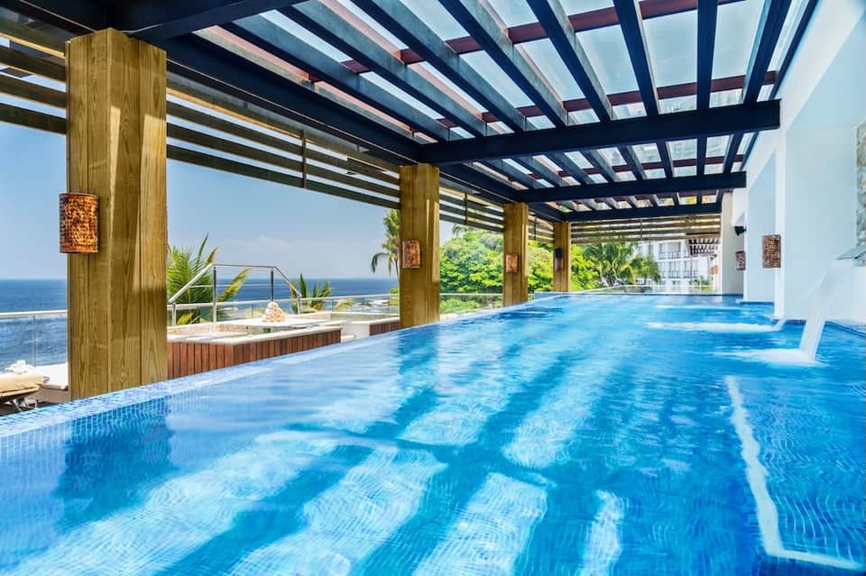 Indoor swimming pool with blue water under wooden pergola overlooking ocean and palm trees
