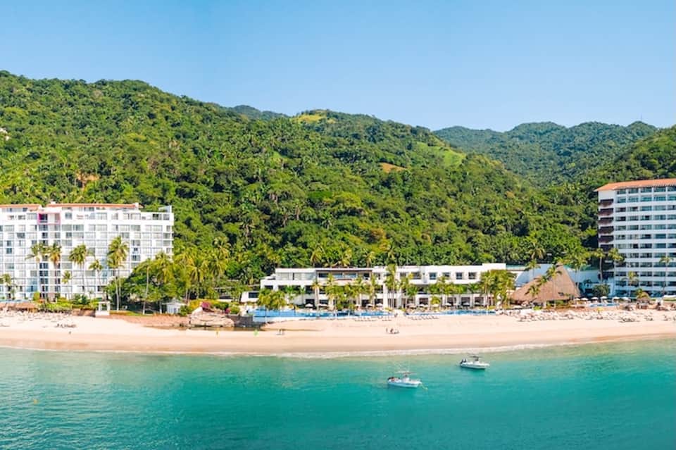 Beachfront resort with white buildings, sandy beach, palm trees, and tropical hillside backdrop