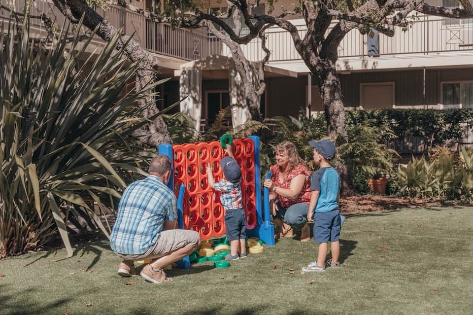 Family courtyard with children playing Connect Four game under mature olive tree with apartment building