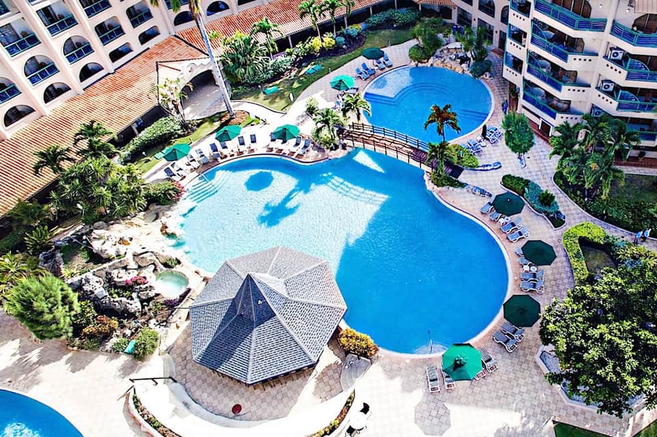 Aerial view of resort with curved blue pools, palm trees, and surrounding apartment buildings