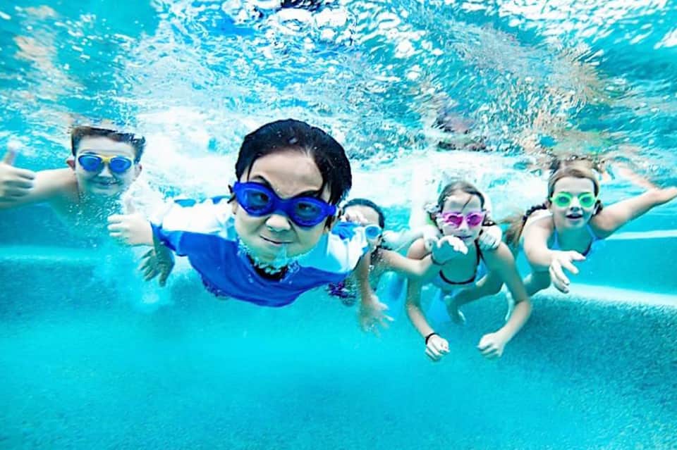 Children swimming underwater wearing colorful goggles in a bright turquoise pool