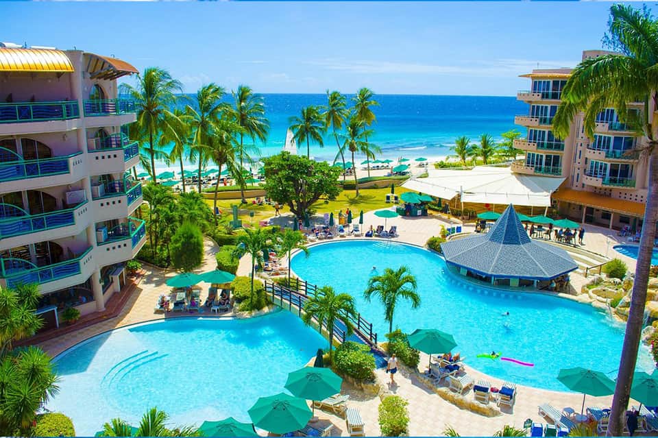 Aerial view of beachfront resort with multiple turquoise pools, palm trees, sandy beaches, and colorful umbrellas