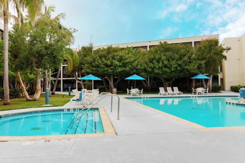 Two connected outdoor pools with lounge chairs, blue umbrellas, mature trees, and building backdrop