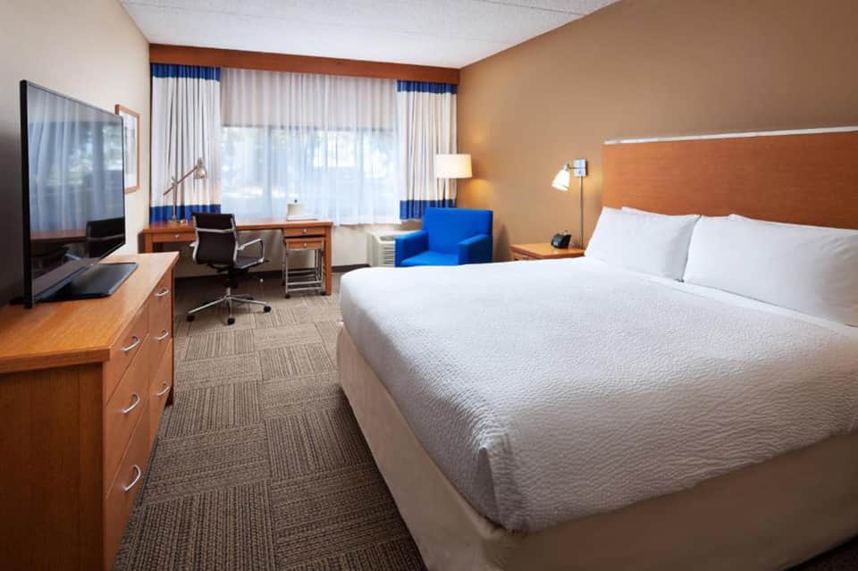 Hotel guestroom with queen bed, work desk, blue accent chair, and large windows with ocean view