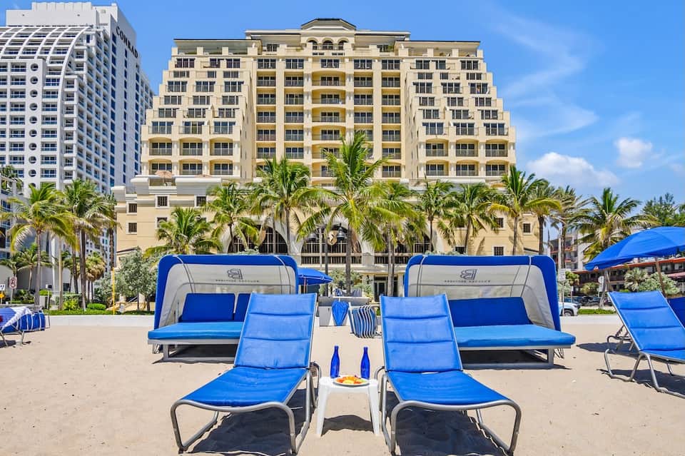 Beach cabanas with blue cushioned lounge chairs in front of multi-story beachfront resort hotel