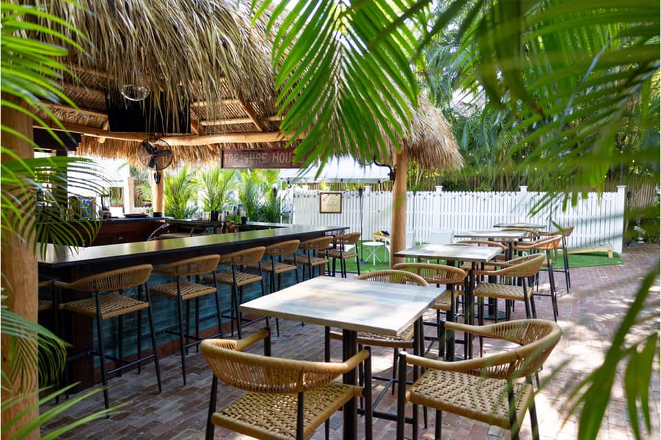 Outdoor dining area with thatched roof, woven chairs, tables, palm fronds, and white picket fence