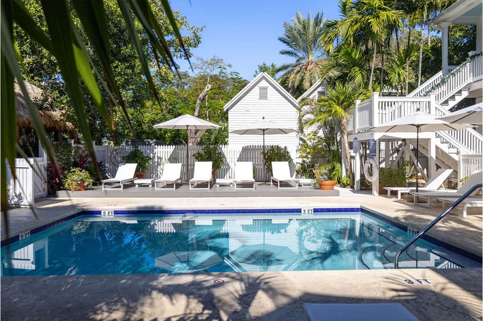 Resort pool surrounded by white lounge chairs, palm trees, and colonial-style white building architecture