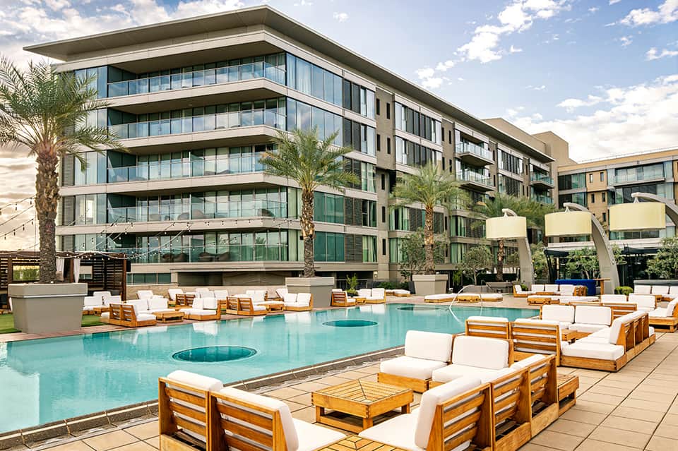 Resort pool with turquoise water, wooden sun loungers with white cushions, and modern building