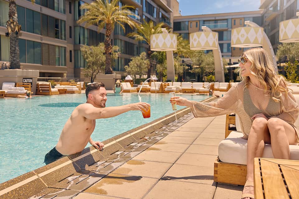 Man and woman toasting drinks at pool edge of luxury resort with lounge chairs and palm trees